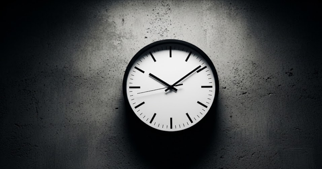Photograph of a single analog wall clock centered against a neutral industrial wall, evenly lit with high contrast and no surrounding activity. The image represents time as the primary operational constraint, emphasizing the narrow decision window in which inventory and execution choices must be made before outcomes become fixed.